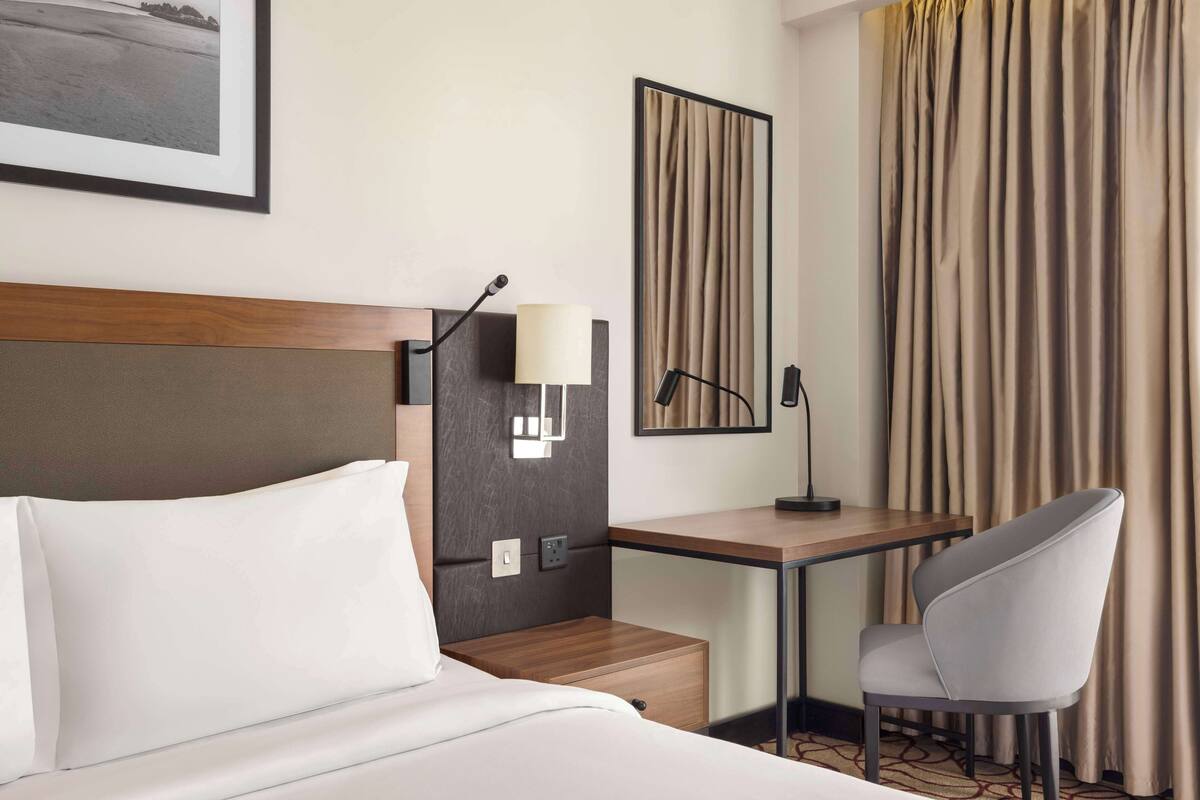 Una habitación de hotel con cama, escritorio, silla, lámpara, espejo y cortinas.