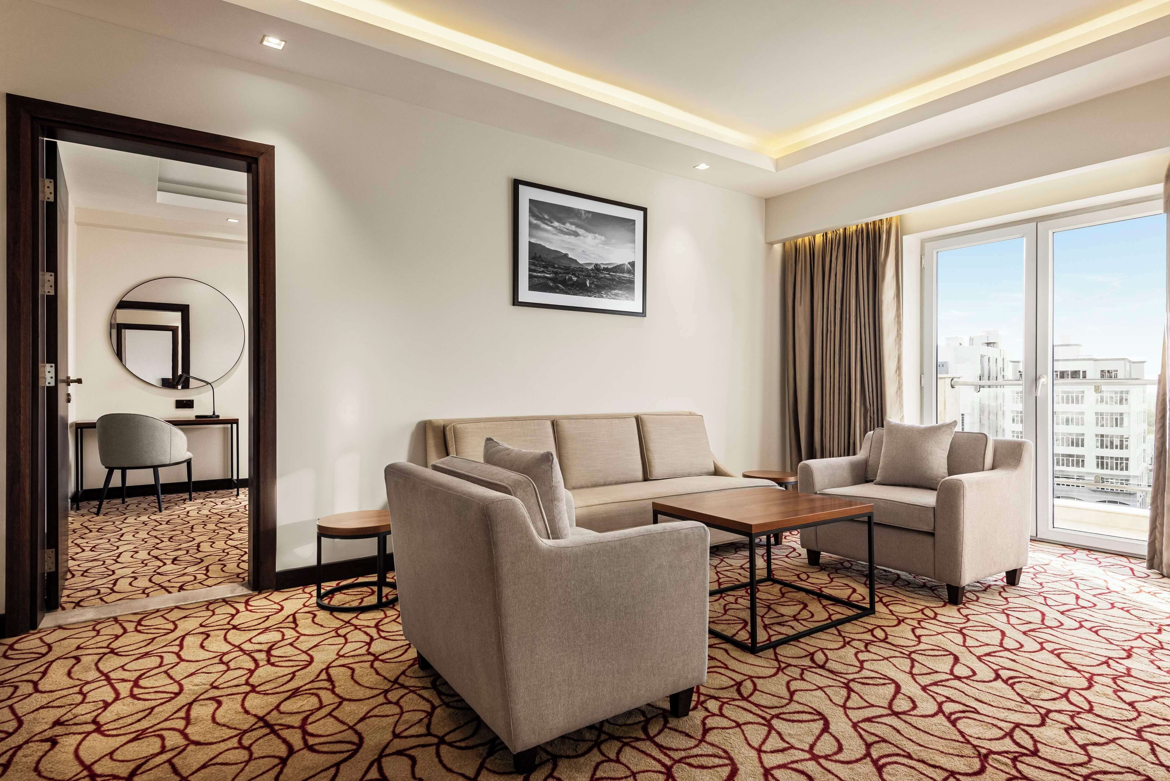 A hotel room with a sofa, armchair, and a small table. There is a large window with curtains, a mirror on the wall, and a framed picture above the sofa.