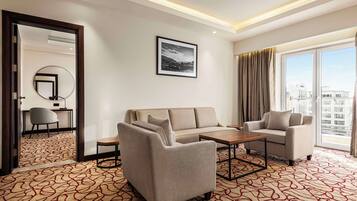 A hotel room with a sofa, armchair, and a small table. There is a large window with curtains, a mirror on the wall, and a framed picture above the sofa.