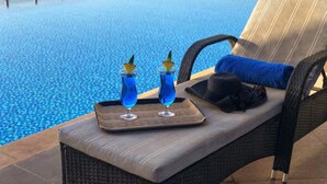 Un área de descanso junto a la piscina, con una silla acolchada, una bandeja con dos bebidas y una toalla azul.