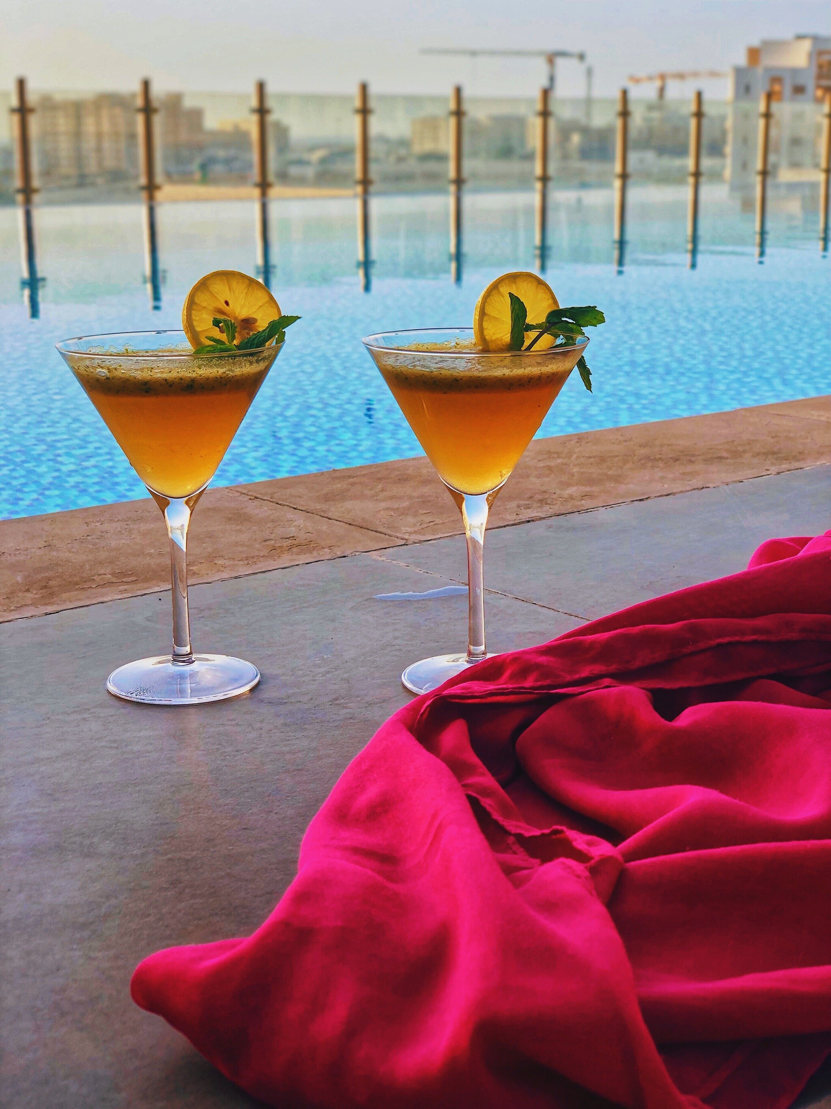 Twee cocktails met garnering op een terras bij een zwembad, met een rode doek en een stadsgezicht op de achtergrond.