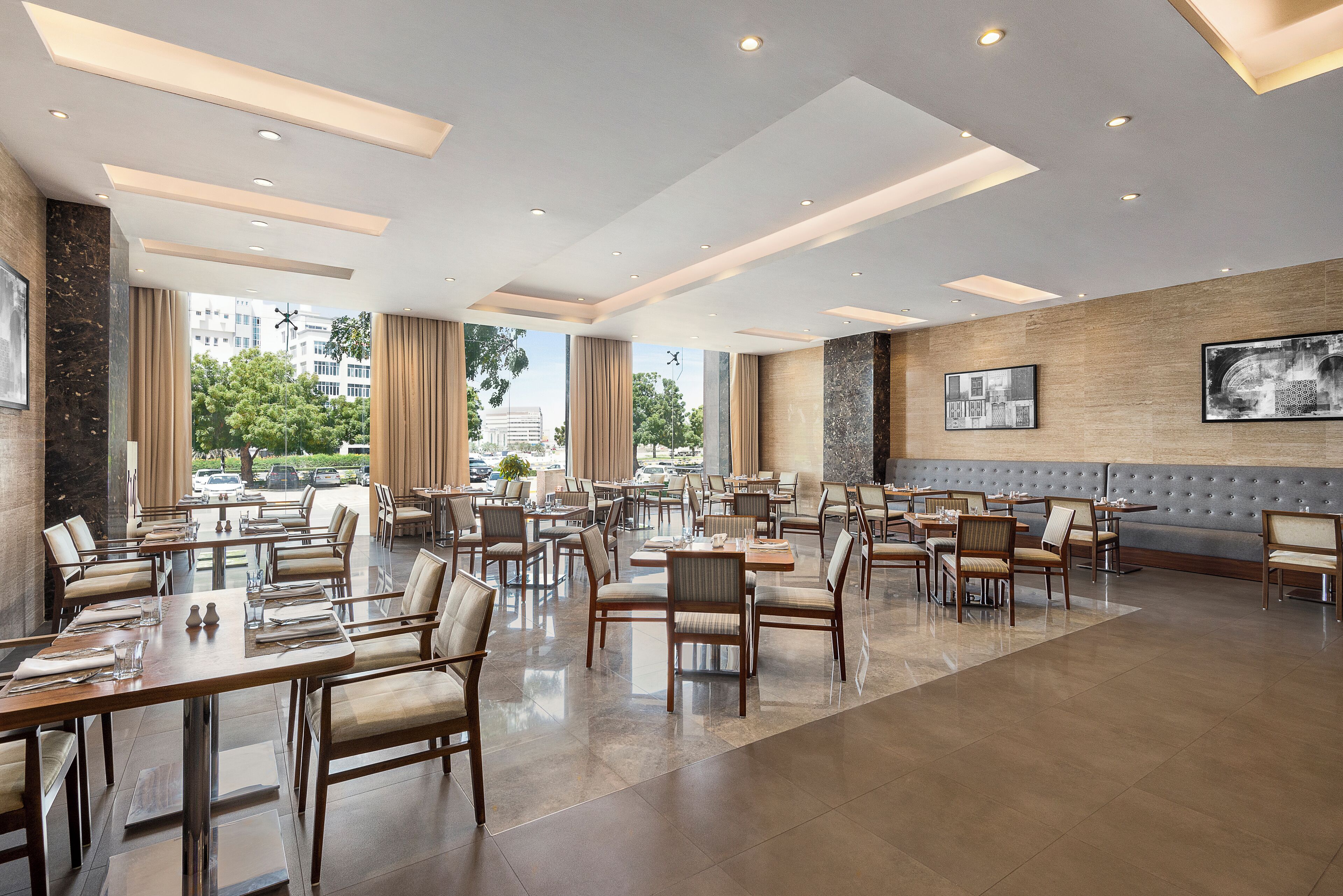 A spacious dining area with multiple tables and chairs, large windows offering a view of the city, and a modern ceiling design.