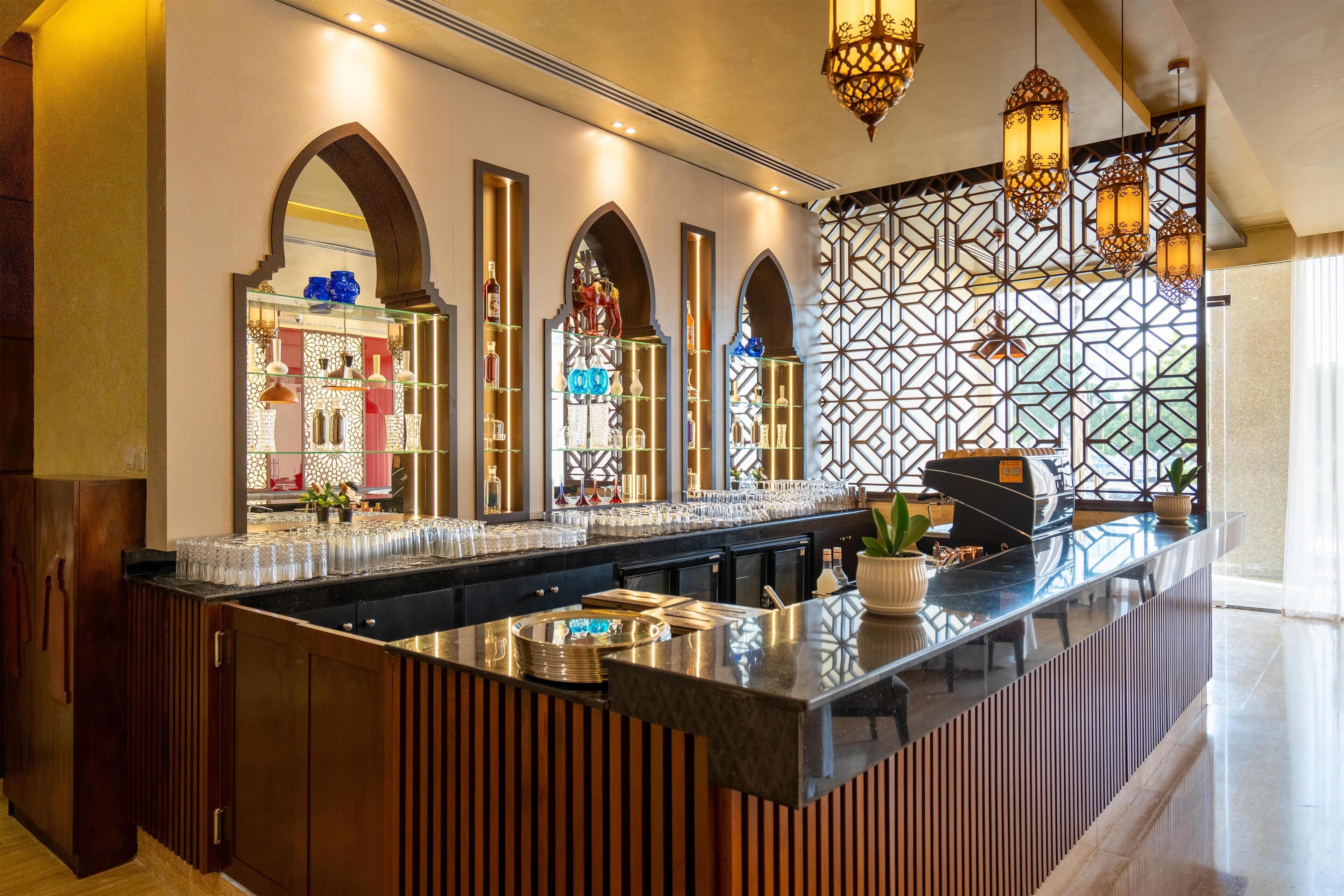A modern bar area with a long counter, multiple shelves with glassware, and decorative lighting.