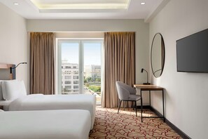 A hotel room with two beds, a desk, a chair, a large mirror, and a view of buildings through the window.
