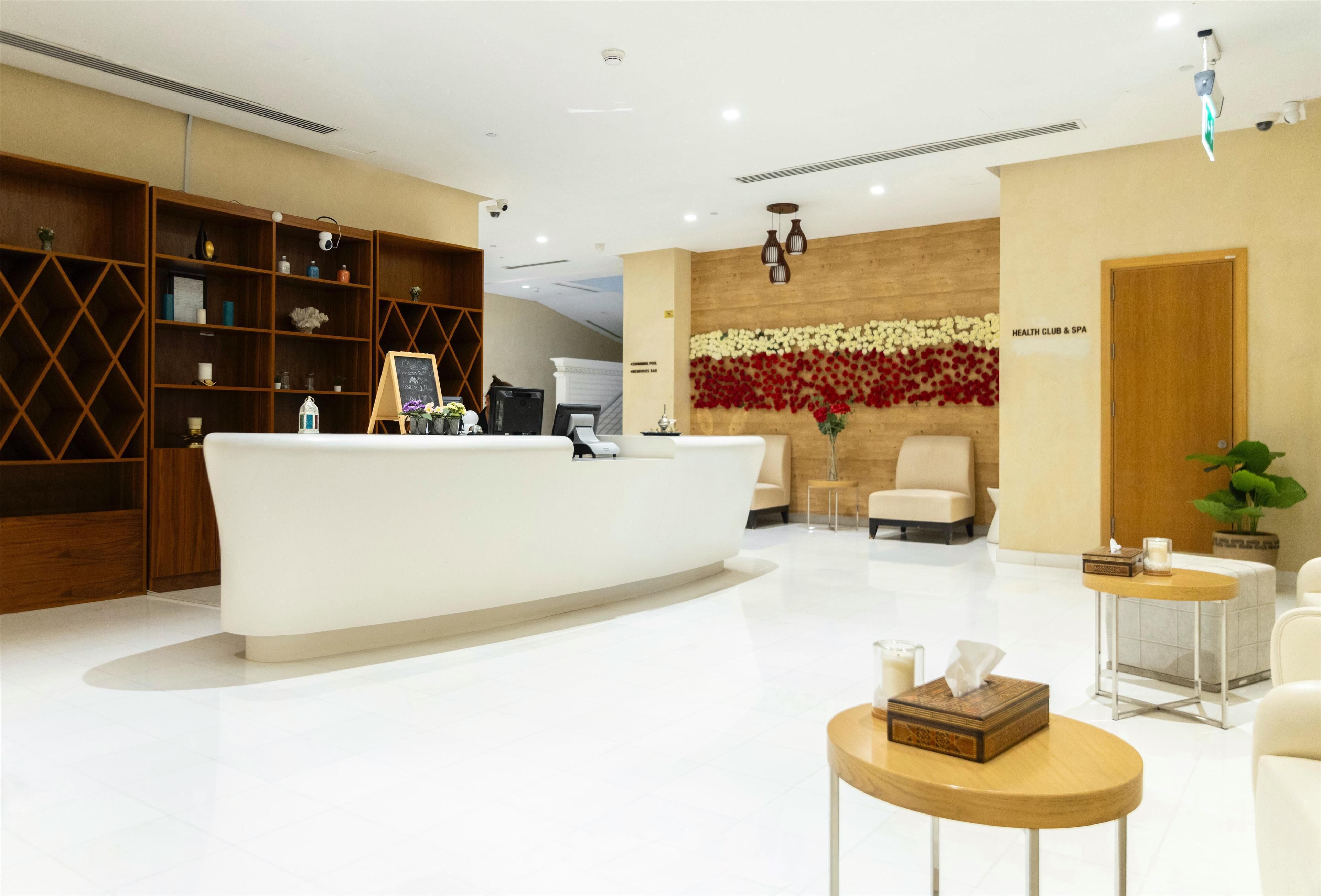 Un área de recepción con un mostrador blanco, estanterías de madera, una pared decorada con flores rojas y blancas, y un letrero que dice "HEALTH CLUB & SPA".