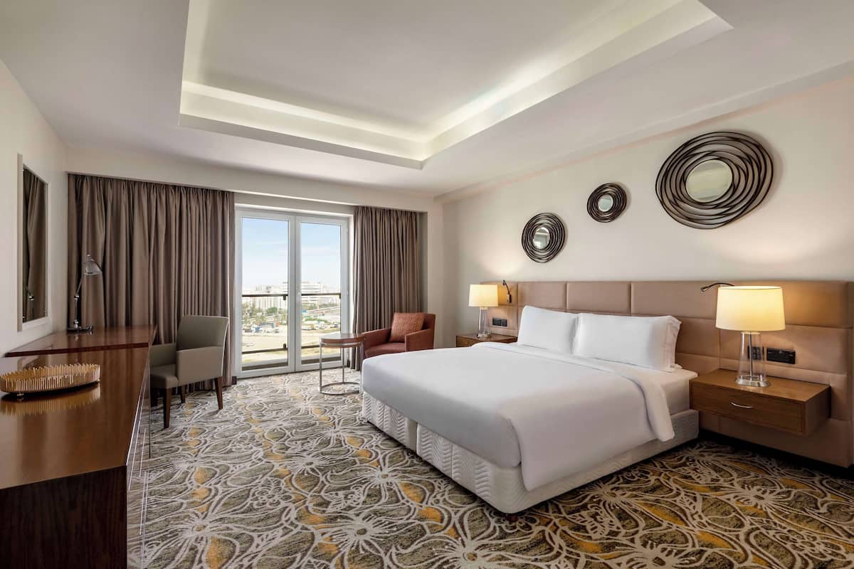 Habitación de hotel con una cama grande, un escritorio, una silla, una mesita de noche con lámparas y vistas a la ciudad desde la ventana.
