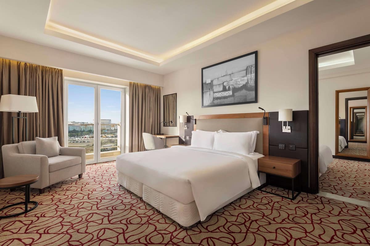 Habitación de hotel con una cama grande, un sofá, un escritorio y una silla. Cuenta con un ventanal con cortinas, un cuadro en la pared y un espejo.
