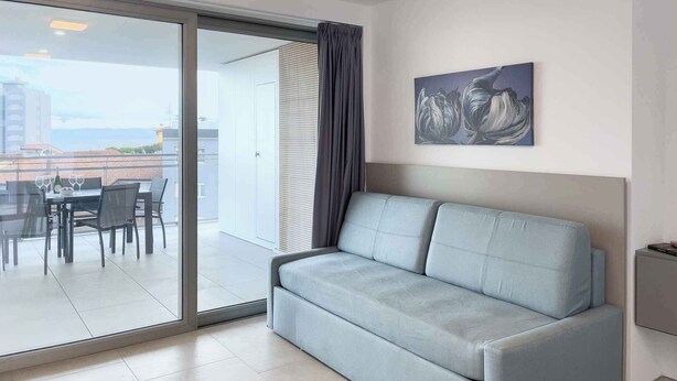 Una sala de estar moderna con un sofá gris, un comedor con sillas y una mesa, y un ventanal con vistas a edificios y una masa de agua.