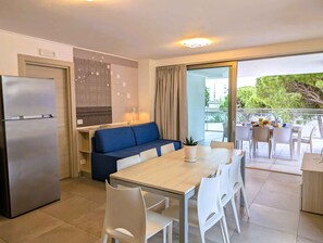 Eine moderne Küche mit integriertem Kühlschrank, eine Essecke mit Holztisch und weißen Stühlen, ein blaues Sofa und ein Balkon mit Blick auf Bäume und Gebäude.