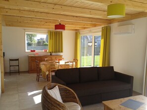 Ein gemütliches Wohnzimmer mit einer Couch, einem Korbstuhl, einem Esstisch mit Stühlen und einer Küchenzeile mit Fenster und Vorhängen.