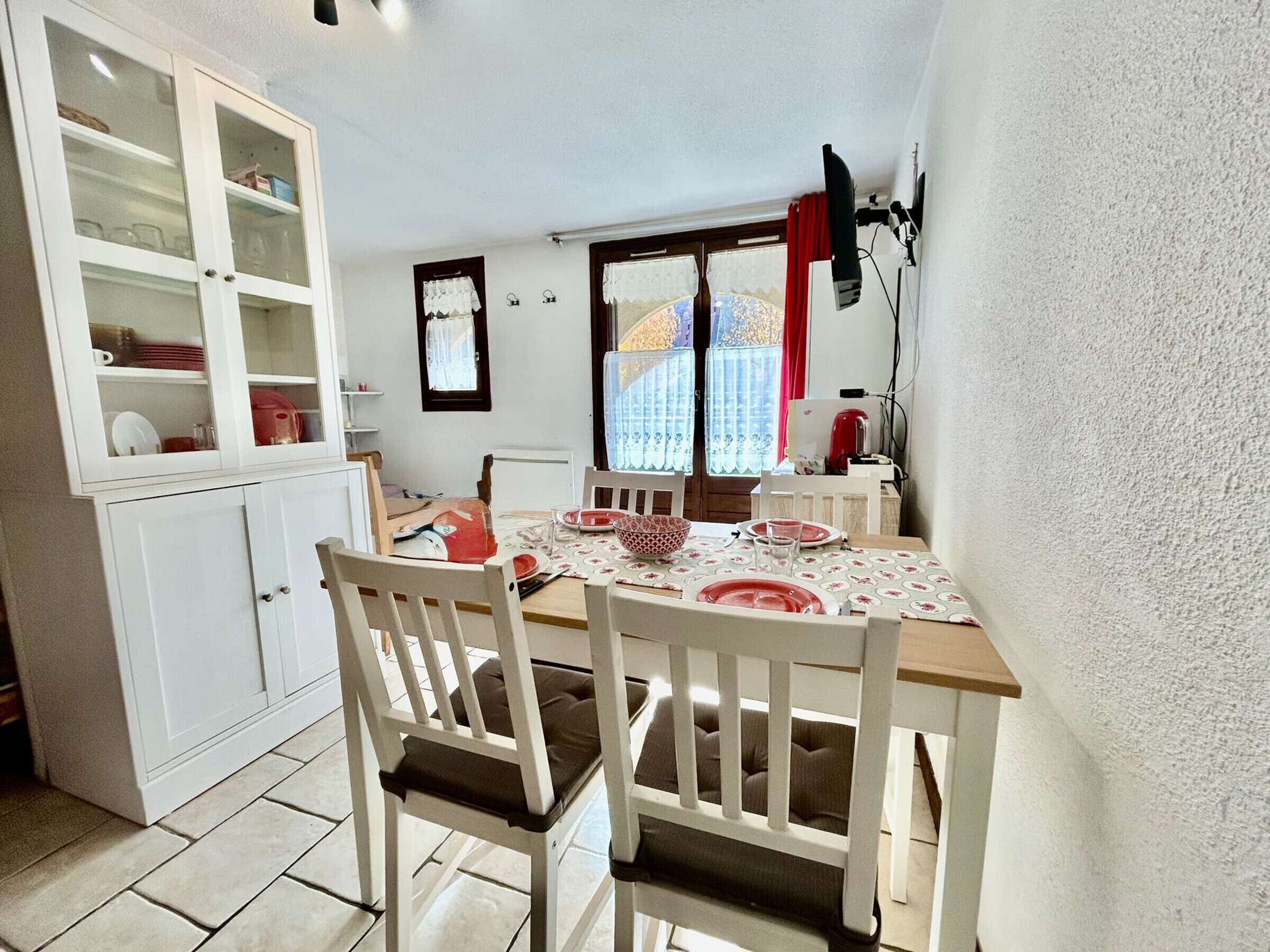 Een eetgedeelte met een houten tafel voor vier personen, witte stoelen, een kast met glazen deuren en een raam met gordijnen.