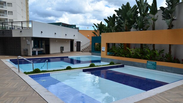Une piscine dont le fond est recouvert de carreaux bleus et blancs, entourée d’une terrasse en bois et bordée d’un bâtiment à la façade blanche et au toit végétalisé.