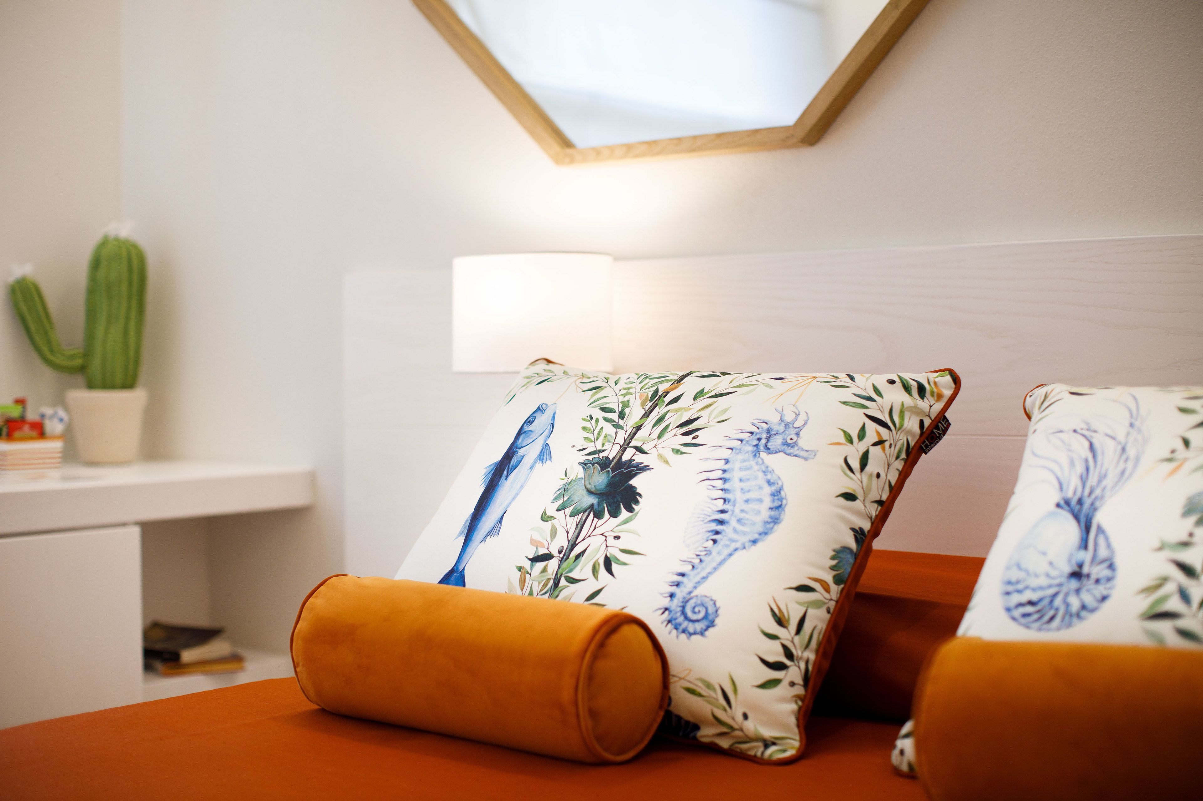 Quarto de hotel bem arrumado com cama, abajur, um cacto e uma pequena estante com livros e uma planta.