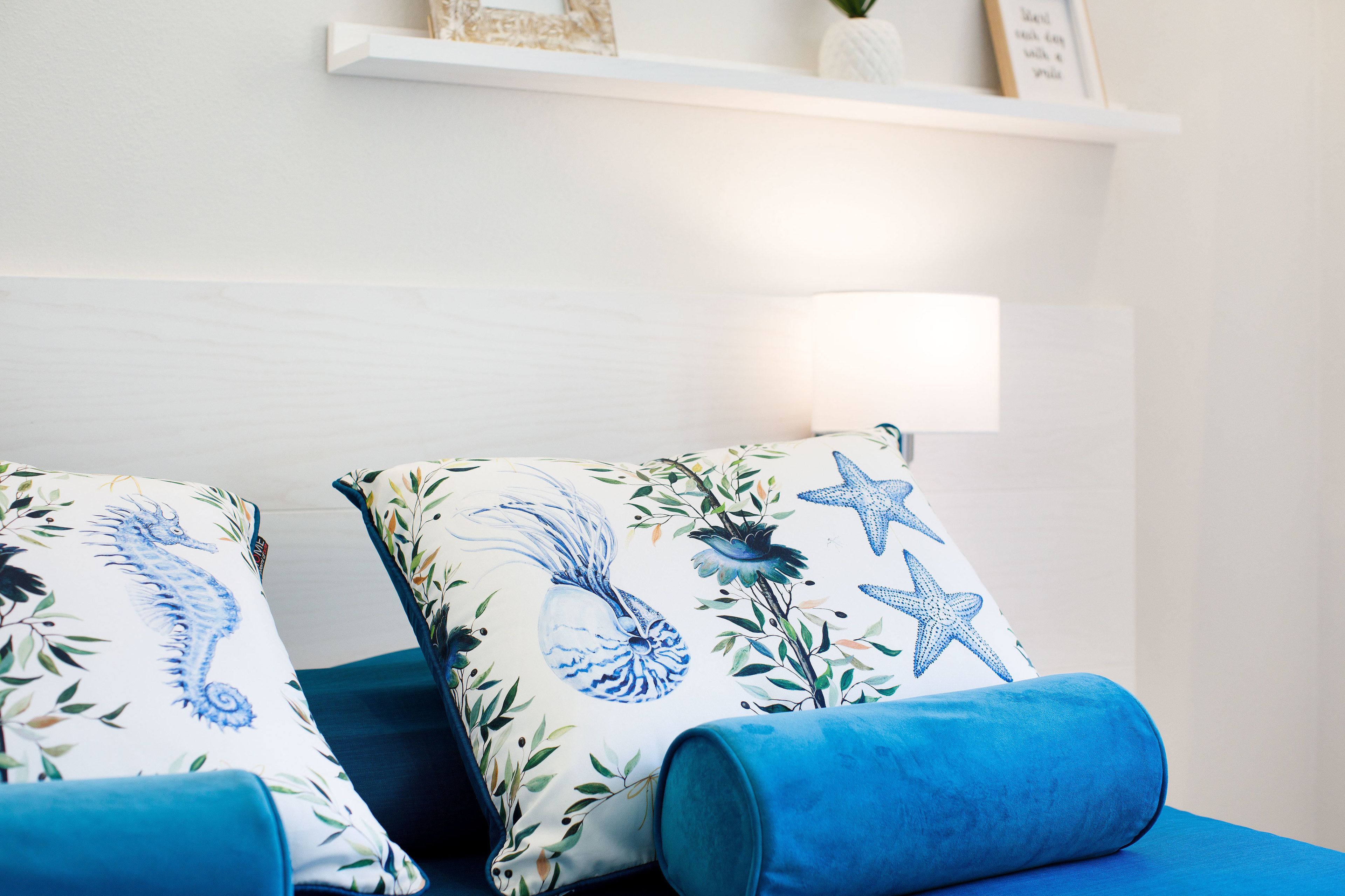 Ein Schlafzimmer mit einem Bett, zwei Kissen mit Seepferdchen- und Sternfisch-Motiv, einer blauen Decke und einer weißen Wand mit einem Regal über dem Bett.