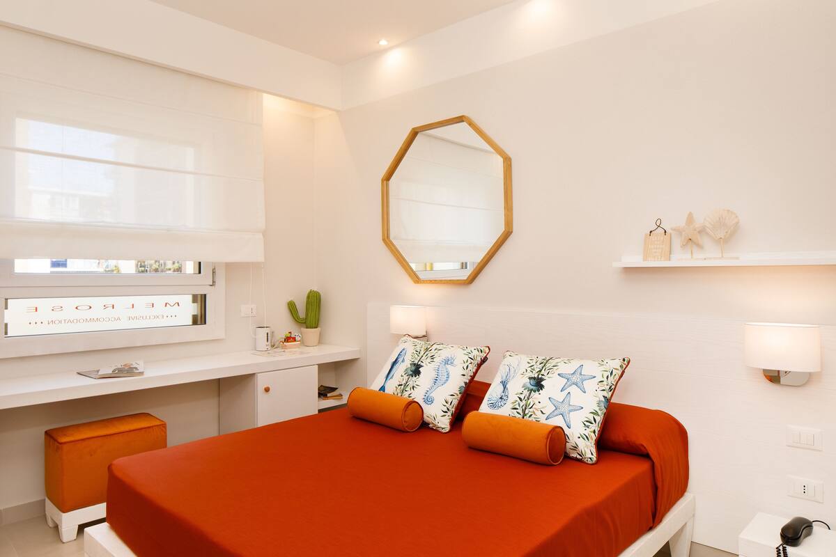 Een moderne hotelkamer met een groot bed, een bureau met een computer, een raam met zonwering, een spiegel en een kleine cactus.