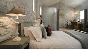 Een slaapkamer met een stenen muur, een bed met witte beddengoed, twee bedlampjes en een badkamer die door een open deur zichtbaar is.