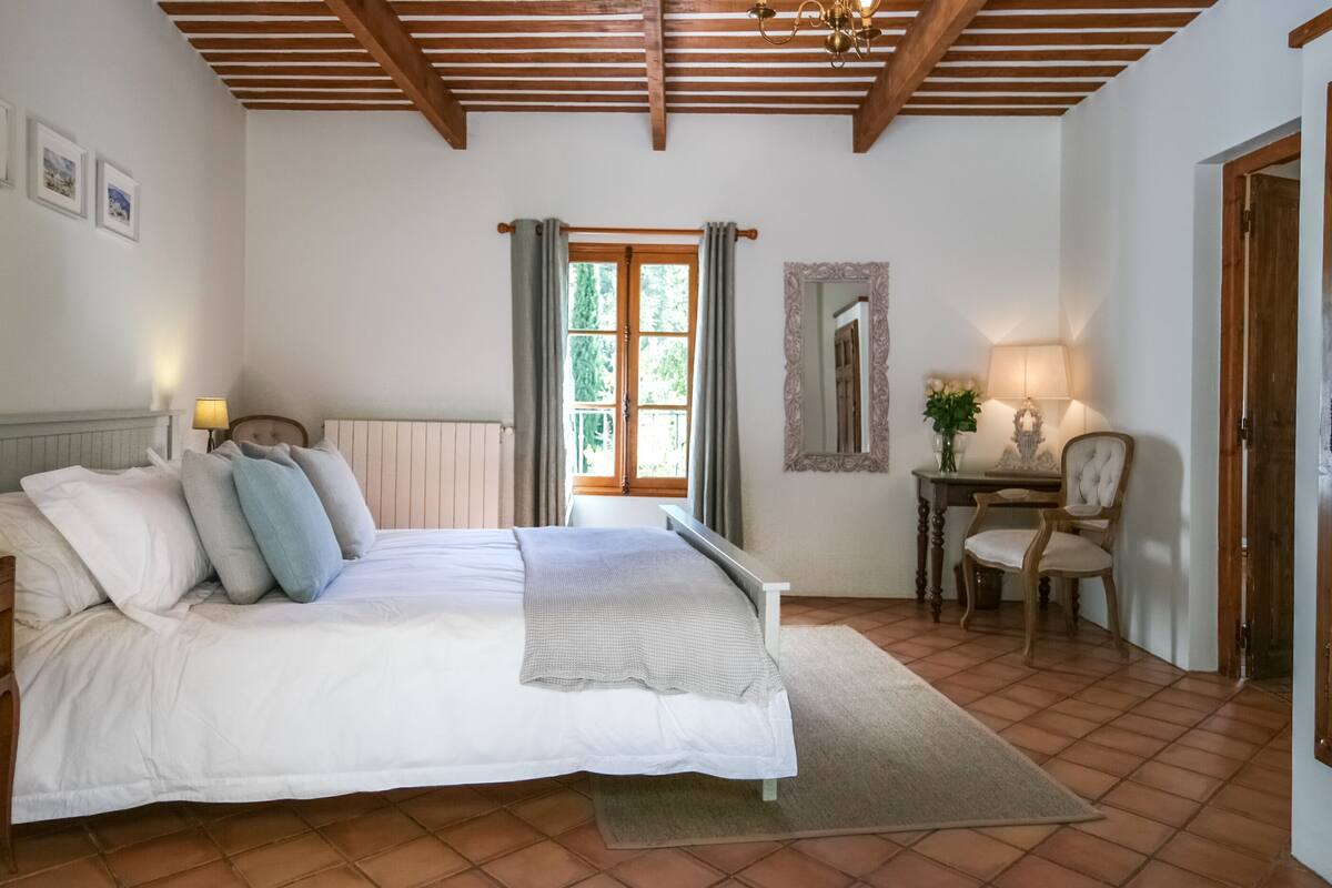 Un dormitorio con cama, mesitas de noche, un escritorio, una silla, un espejo y una ventana con cortinas.