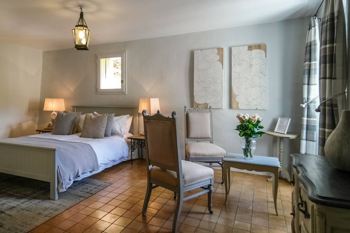 Un dormitorio con una cama, dos mesitas de noche, una silla, una mesita pequeña y un jarrón con flores.