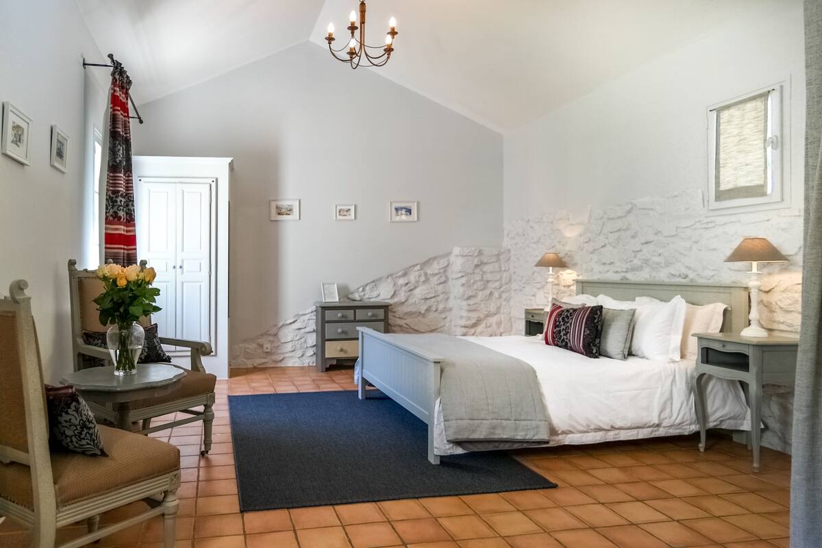 Un dormitorio con cama, mesitas de noche, una silla, un escritorio, una araña y una pared de piedra.