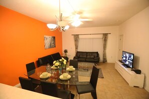Sala de estar moderna com área de jantar, apresentando uma mesa de vidro, cadeiras pretas, um sofá e uma televisão.