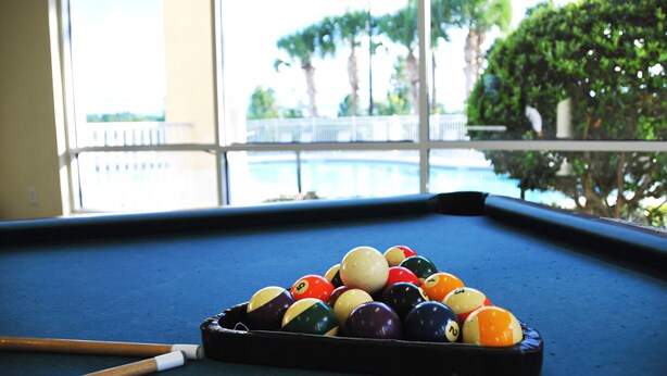 A pool table with balls and a cue stick in a room with large windows overlooking a pool.