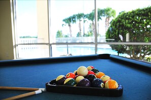 Een pooltafel met ballen en een keu in een ruimte met grote ramen met uitzicht op een zwembad.