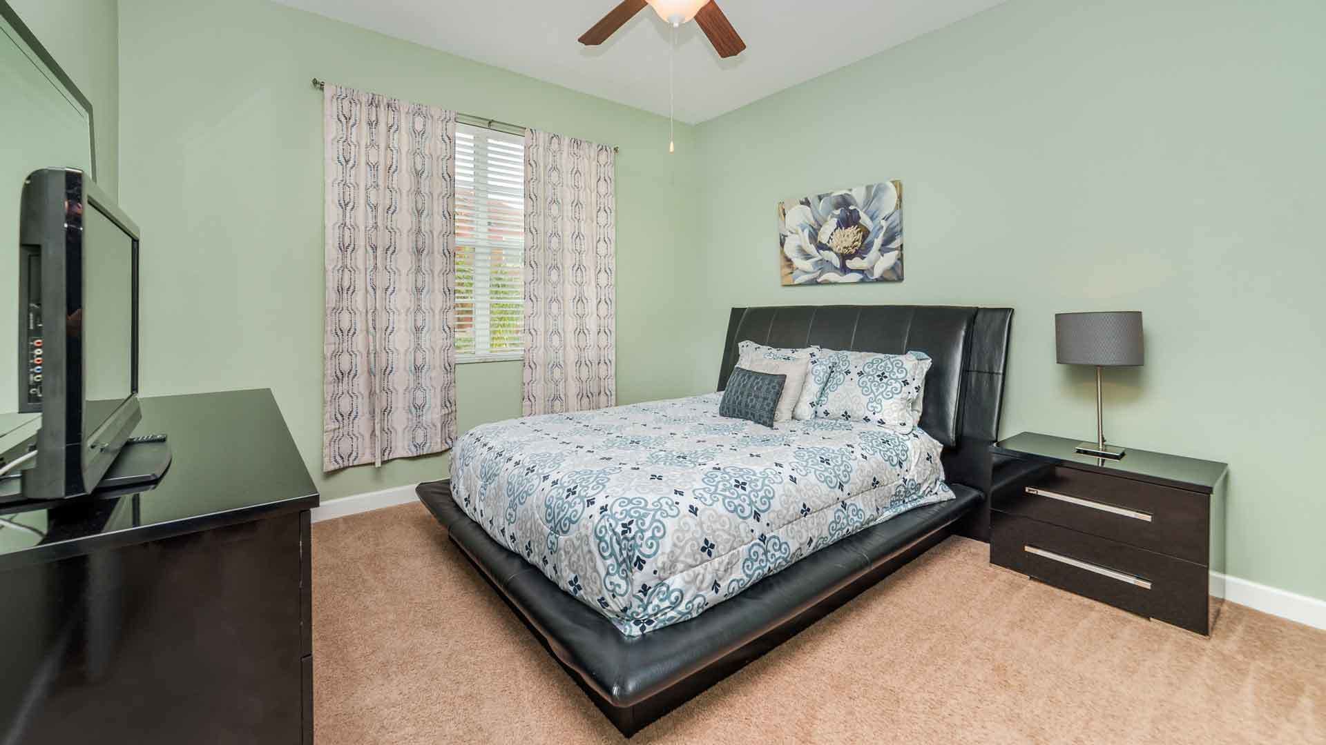 Een slaapkamer met een bed, een bureau, een stoel, een nachtkastje, een lamp en een plafondventilator.