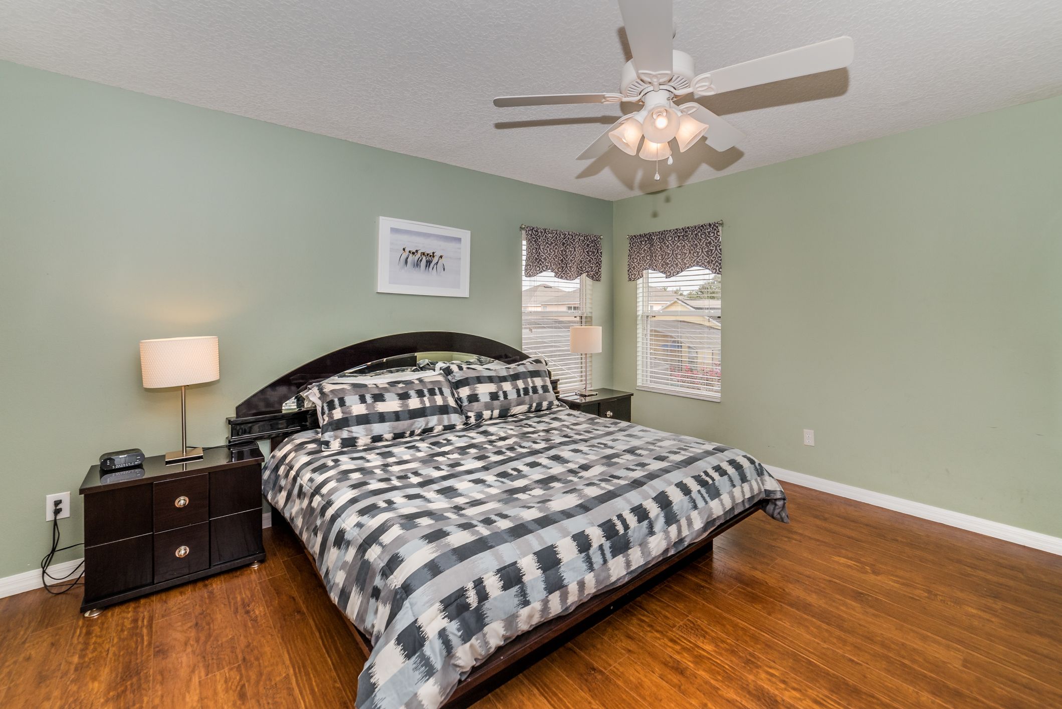 A bedroom with a bed, nightstands, a ceiling fan, and windows with patterned curtains.