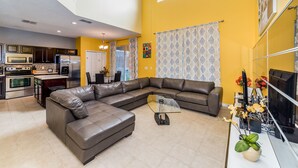A modern living room with a sectional sofa, a glass coffee table, and a kitchen with stainless steel appliances.