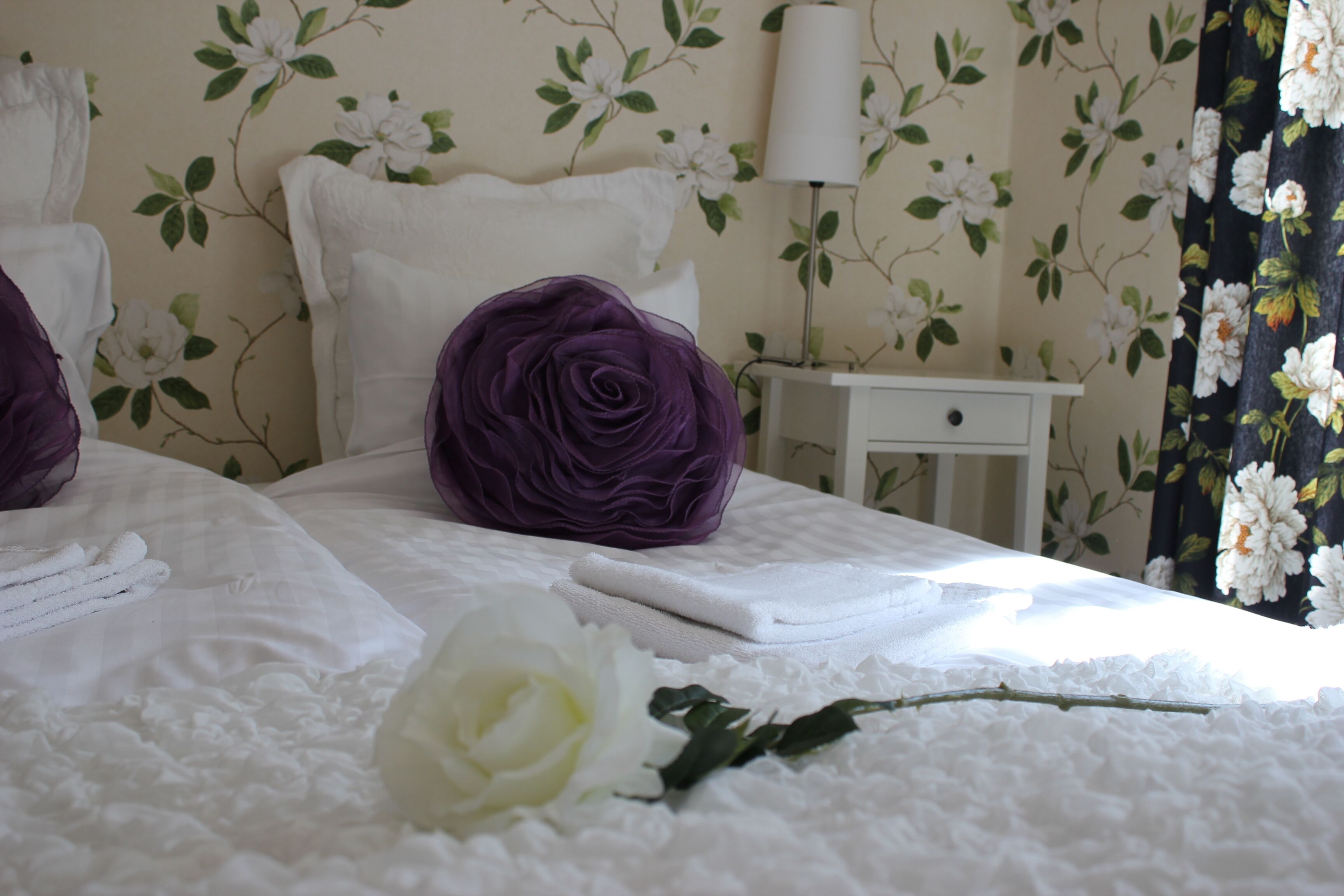 Una cama impecablemente hecha con sábanas blancas, un cojín decorativo morado y una rosa blanca.