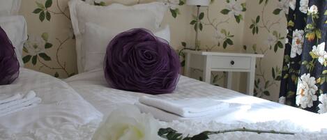 Una cama impecablemente hecha con sábanas blancas, un cojín decorativo morado y una rosa blanca.