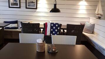 Un comedor con una mesa de madera oscura, sillas de mimbre blanco y un banco con cojines a rayas. Hay cuadros en la pared y una pequeña decoración con forma de velero.
