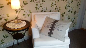 A white armchair with a cushion that reads "Friends of Notte-D'Ami-Champs," a small round table with a lace doily, and a floor lamp.