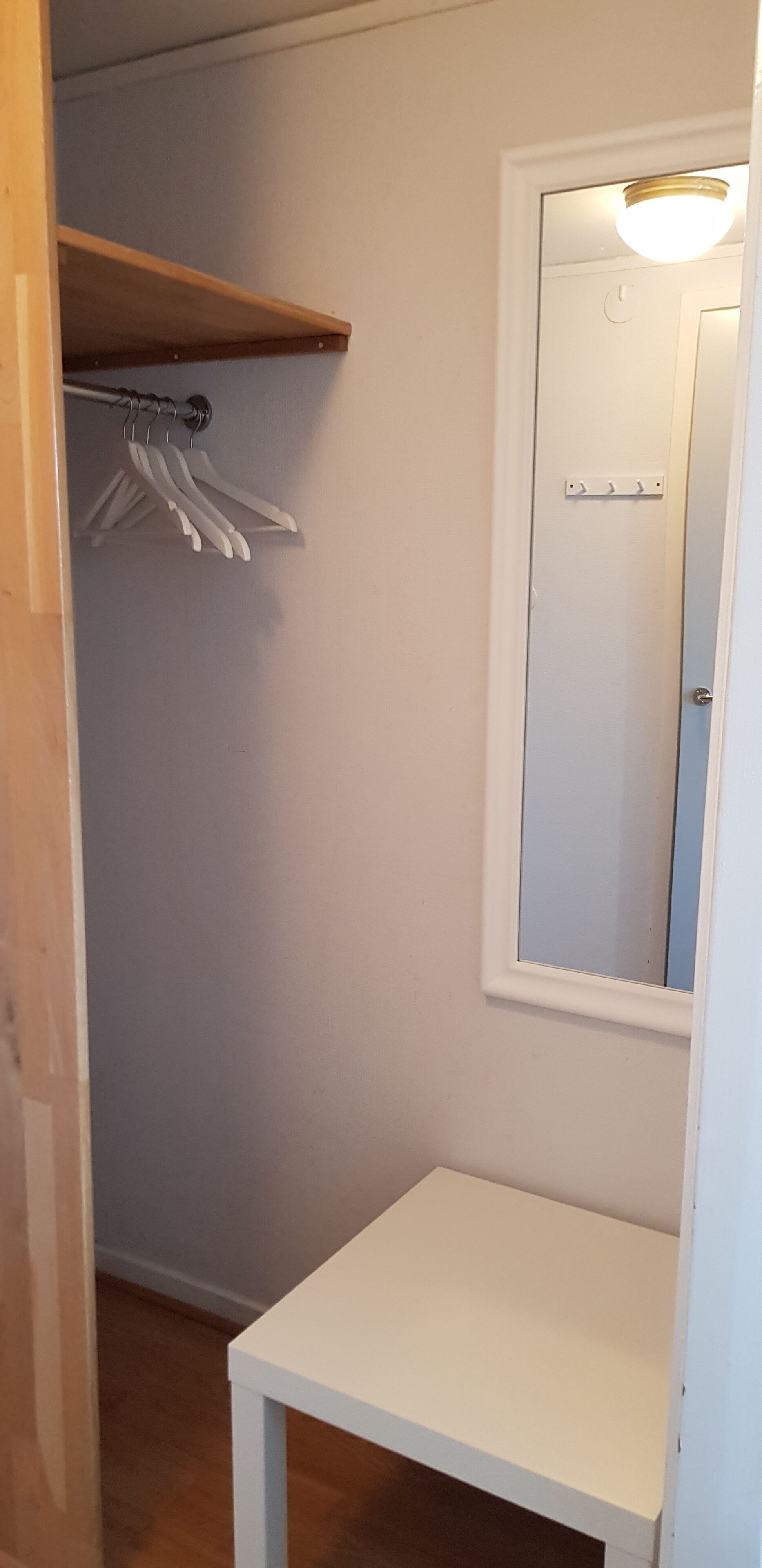A small, white room with a wooden shelf, a mirror, and a white table.