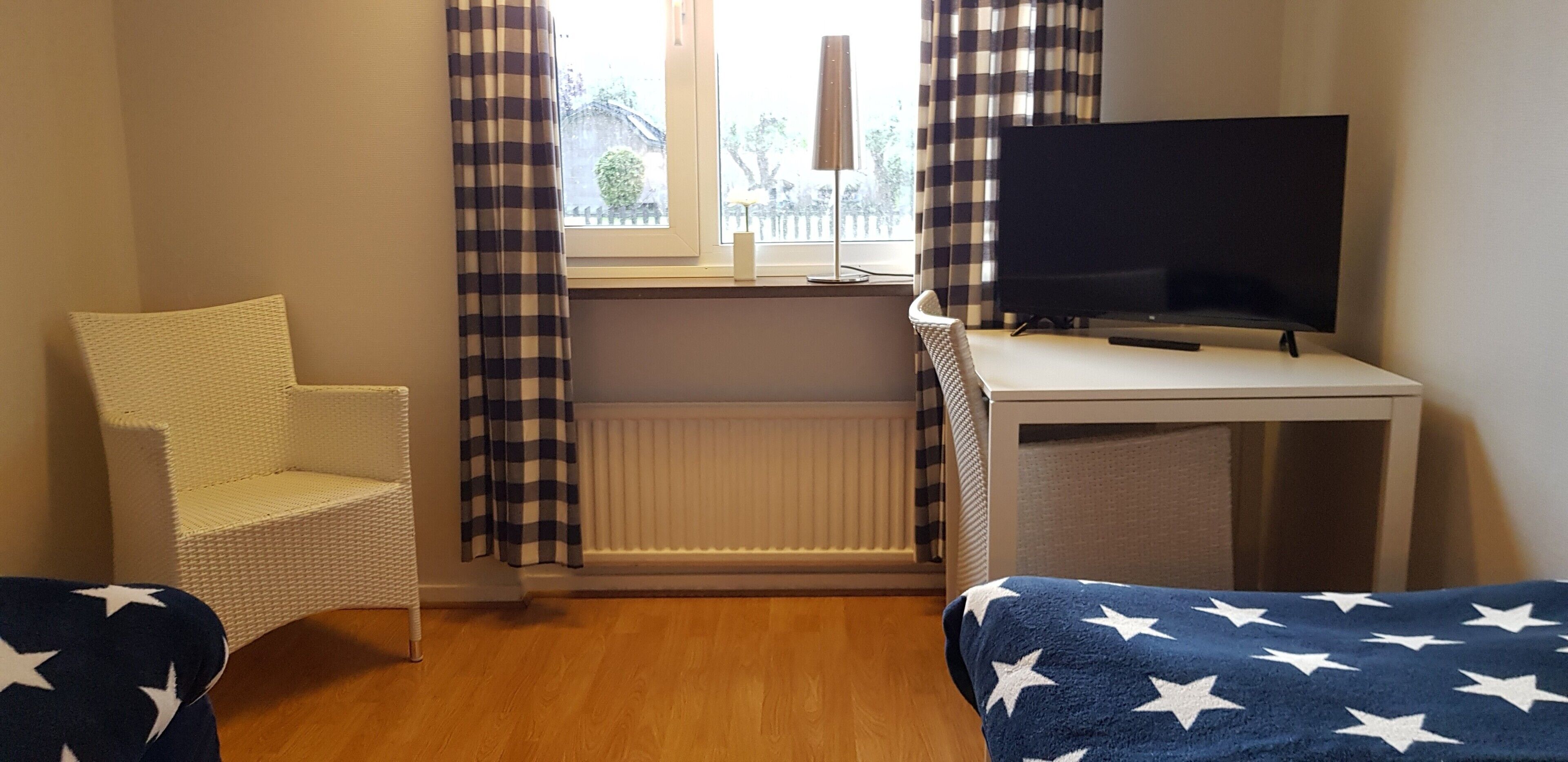 A room with a white chair, a bed with a blue blanket, a TV on a stand, and a window with curtains.