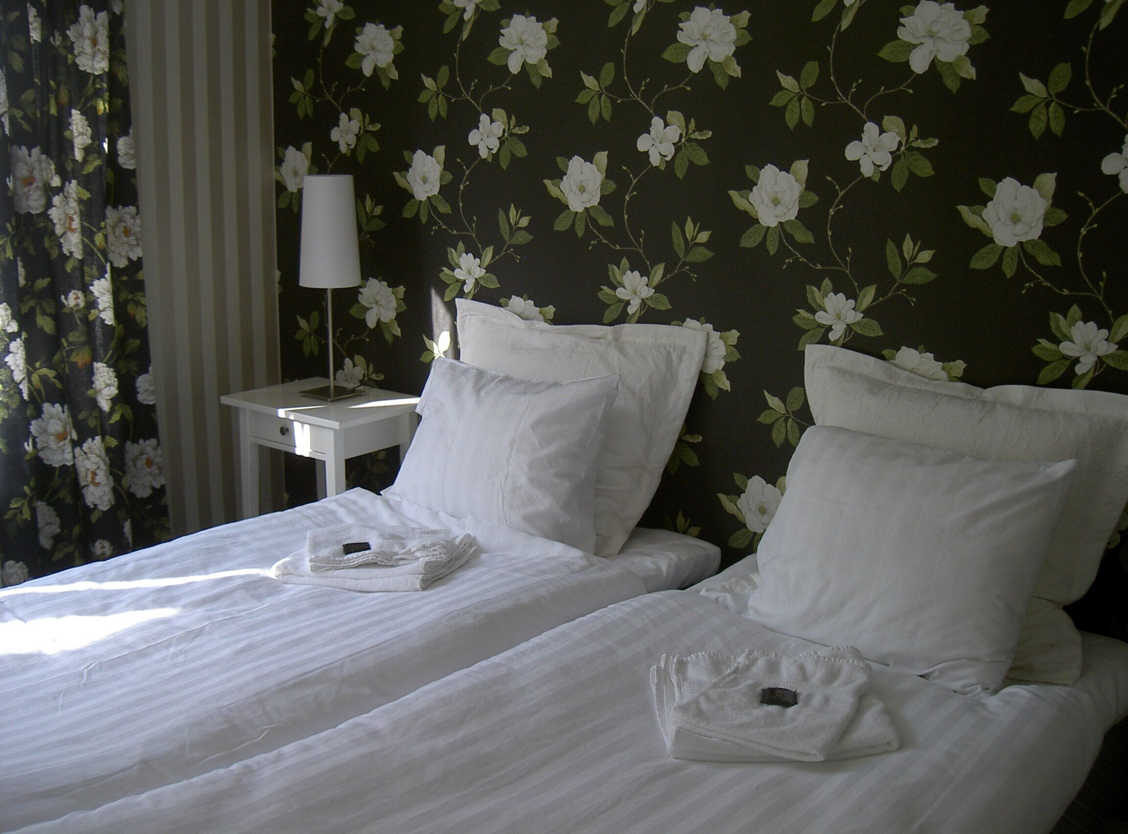 A hotel room with two beds, white linens, a nightstand with a lamp, and floral wallpaper.