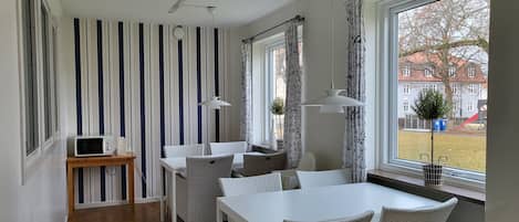 A modern kitchen with striped wallpaper, a white dining table, wicker chairs, a microwave, and large windows with curtains.