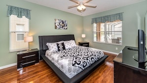 A bedroom with a bed, a ceiling fan, a nightstand, a TV, and two windows with blinds. - Emerald Island #2 - 7 Bed 5 Baths Villa (Kissimmee)