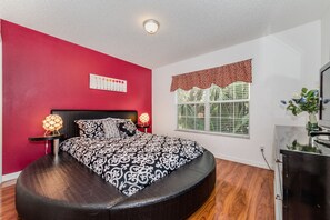Ein Schlafzimmer mit roter Wand, einem Bett mit gemustertem Überwurf, einer Nachttischlampe, einem Fenster mit blickdichtem Vorhang und einem Fernseher auf einem Schrank.
