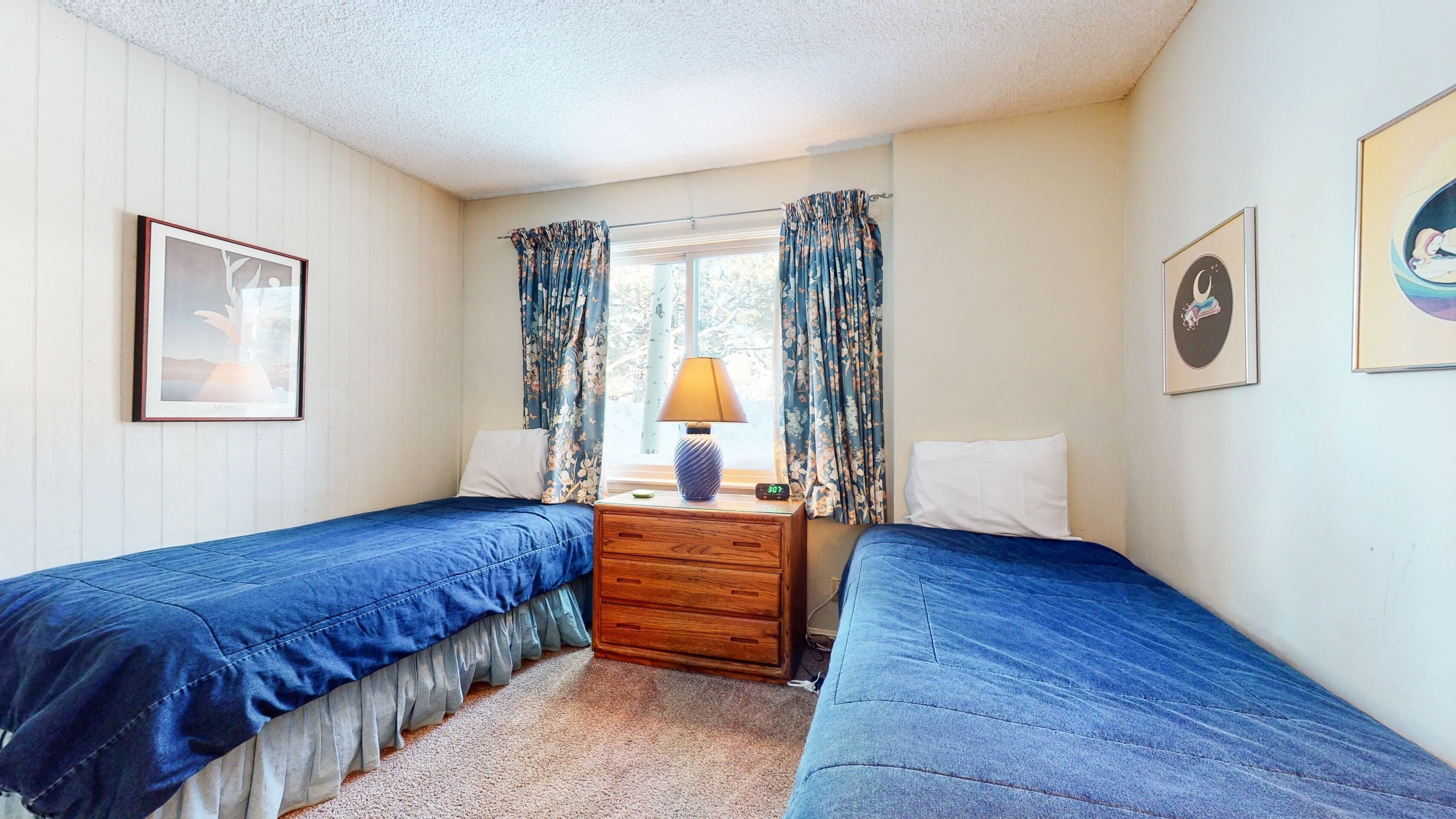 A small, cozy bedroom with two single beds, a wooden nightstand with a lamp, and a window with floral curtains.