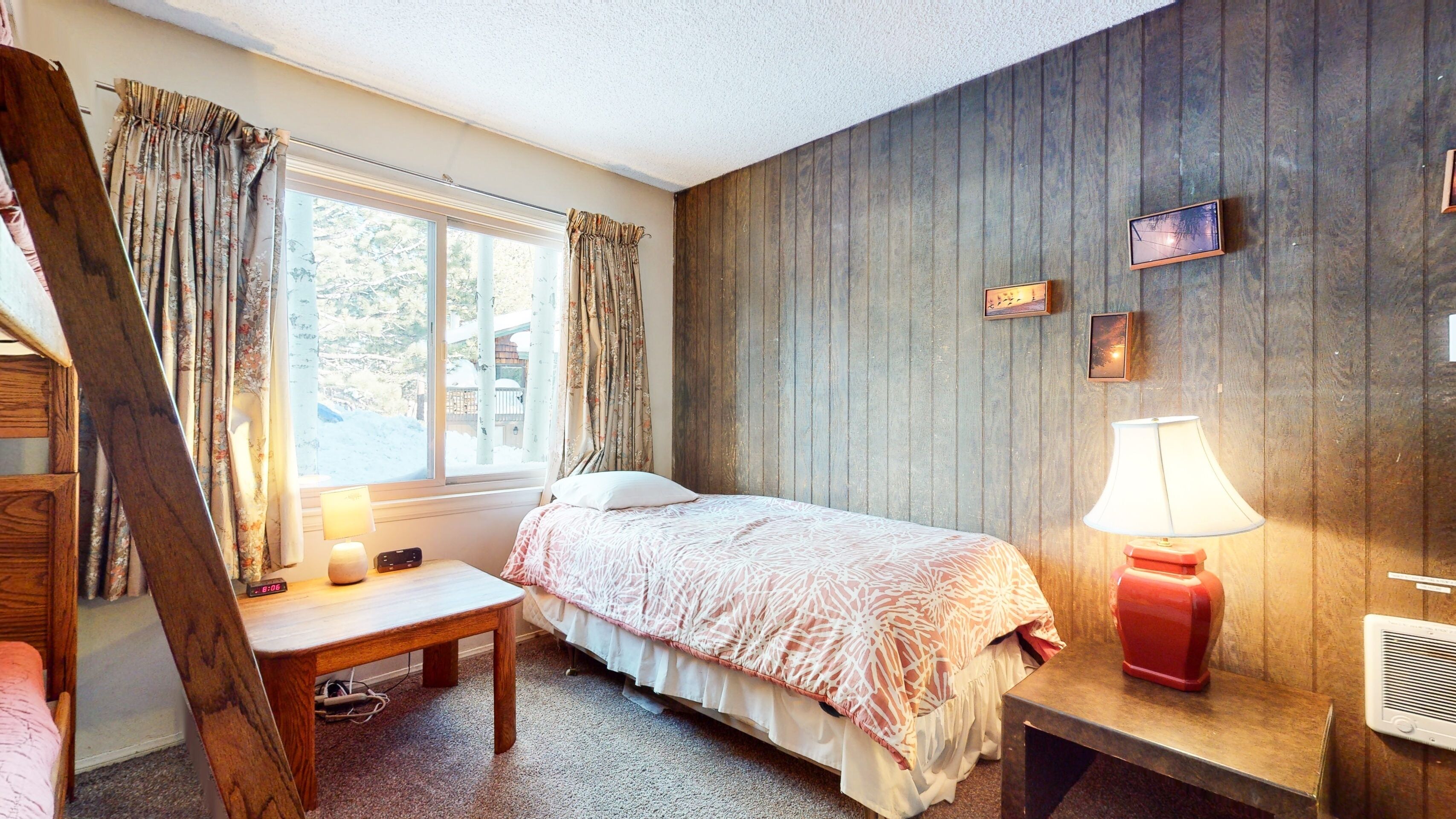 Ein Schlafzimmer mit einem Bett, einem Nachttisch mit Lampe, einer Holzwand und einem Fenster mit Vorhängen.