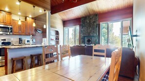 A cozy cabin interior with a wooden dining table, chairs, a stone fireplace, and a large window offering a view of the outdoors.