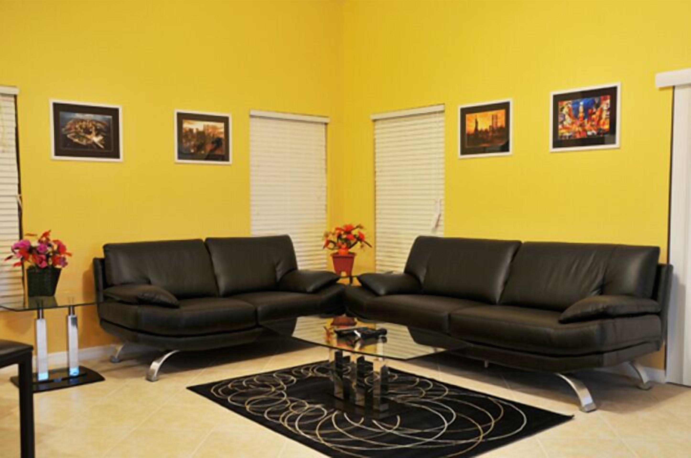 A living room with two black leather sofas, a glass coffee table, and a yellow wall with framed pictures.