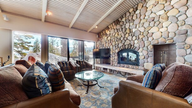 A cozy living room with stone walls, a fireplace, comfortable seating, and a large window offering a view of the outdoors. - Mammoth West 137 Updated Deluxe Condo, Just A Short Walk to Canyon Lodge by RedAwning (Mammoth Lakes)