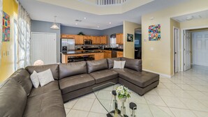 A spacious living room with a large sectional sofa, a glass coffee table, and a kitchen in the background.