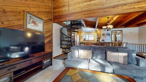 A cozy living room with a large TV, a sofa, and a wooden staircase leading to a kitchen. - Snowflower 26 Remodeled Spacious Condo With Great Complex Amenities by RedAwning (Mammoth Lakes)
