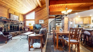 A cozy cabin interior with a stone fireplace, wooden furniture, a TV, and a kitchen area. - Snowflower 26 Remodeled Spacious Condo With Great Complex Amenities by RedAwning (Mammoth Lakes)