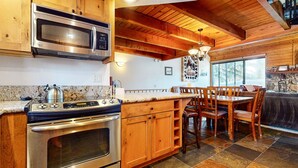A kitchen with wooden cabinets, a stainless steel stove, a microwave, and a dining area with wooden chairs and a table. - Snowflower 26 Remodeled Spacious Condo With Great Complex Amenities by RedAwning (Mammoth Lakes)