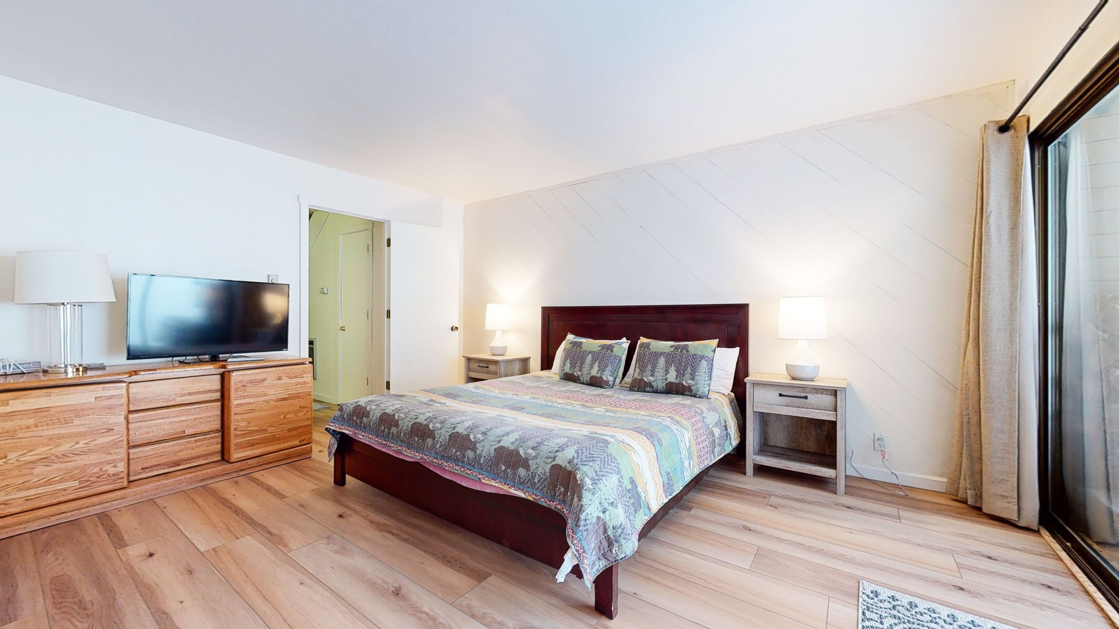 A bedroom with a wooden bed, a television on a dresser, and a large window.