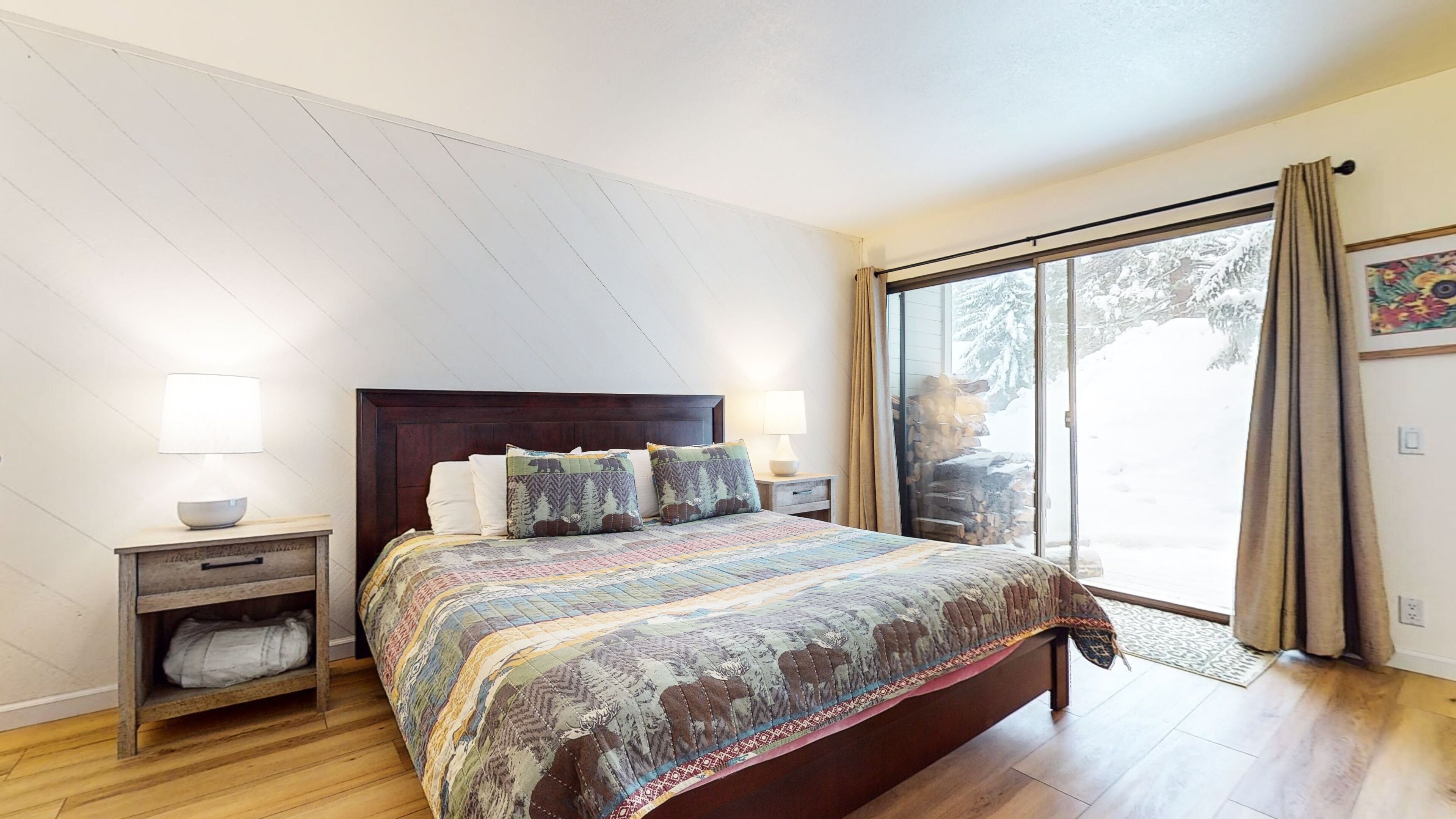 Een slaapkamer met een groot bed, een houten hoofdbord, twee nachtkastjes met lampen, een klein plankje en een schuifdeur van glas die uitkomt op een besneeuwd buitengebied.